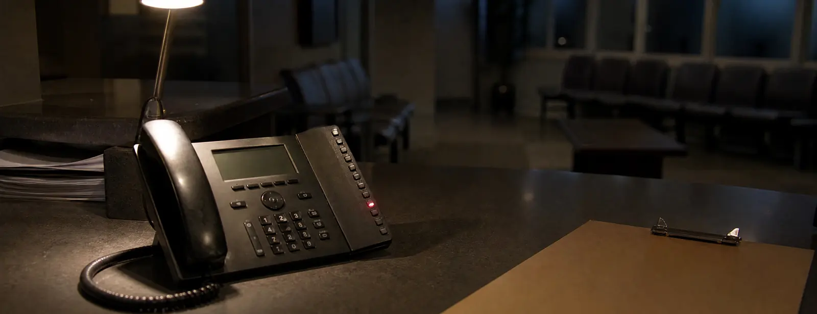 Family medicine front desk after closing: a desk lamp, a clipboard and a desk phone with its line light on, the waiting room dark and empty behind.