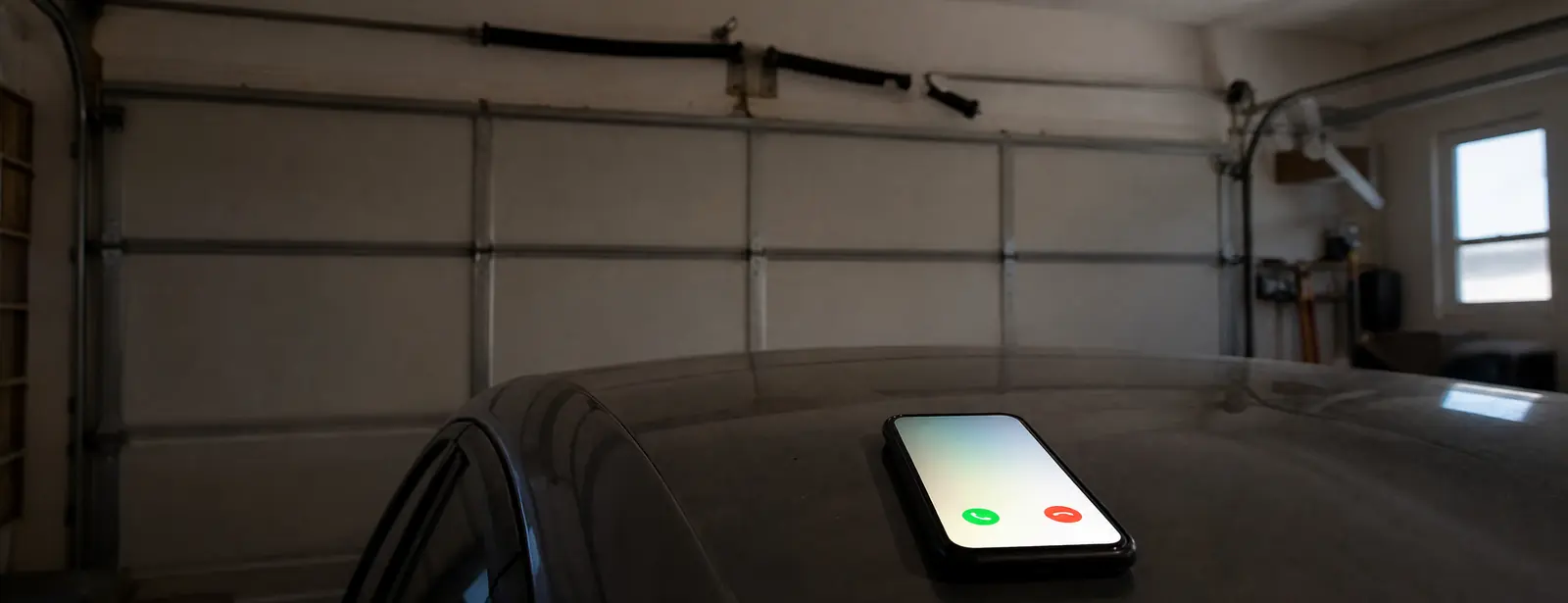 A phone ringing on the roof of a car parked inside a dim garage, under a closed door with a broken spring hanging loose above it.