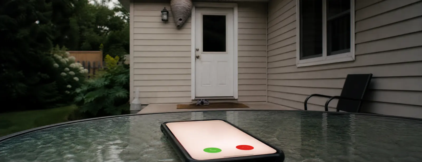 A phone ringing on a glass patio table facing a closed back door, with a large wasp nest under the eave and small sneakers on the mat.