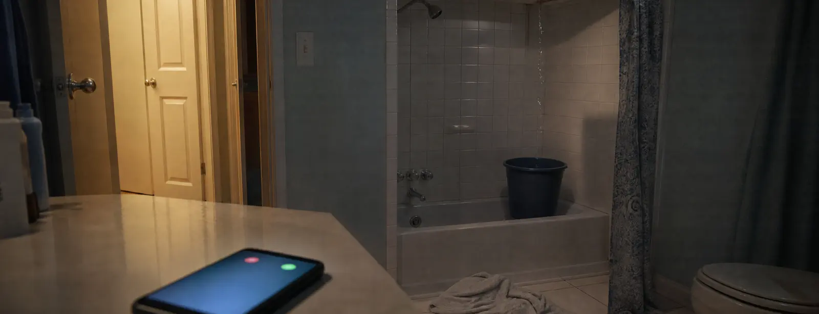 An apartment bathroom at night: water dripping from the ceiling into a bucket in the tub, a towel on the floor and a smartphone glowing on the counter.